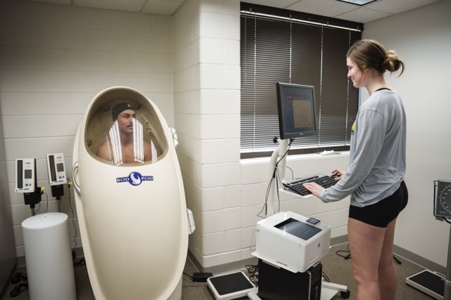 A male subject sits inside a transparent-front Bod Pod body composition chamber while a female technician stands at a computer workstation in a clinical lab, preparing or monitoring the body composition test.