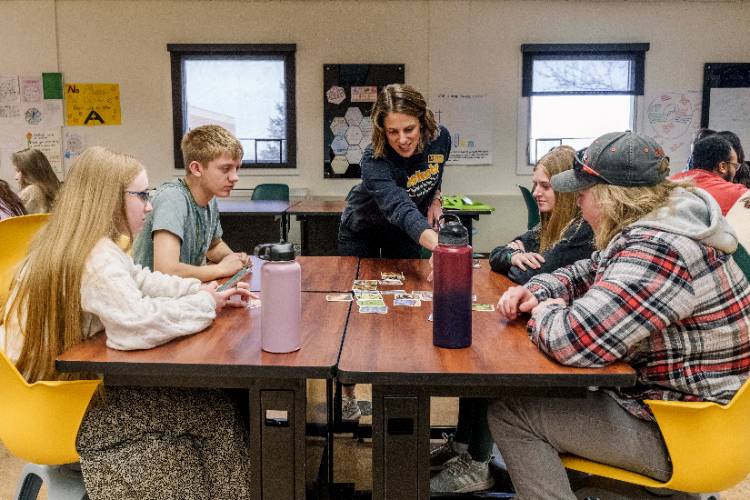 A smiling instructor guides four college-age students playing a card-based learning game around joined tables in a bright classroom with posters and large windows.