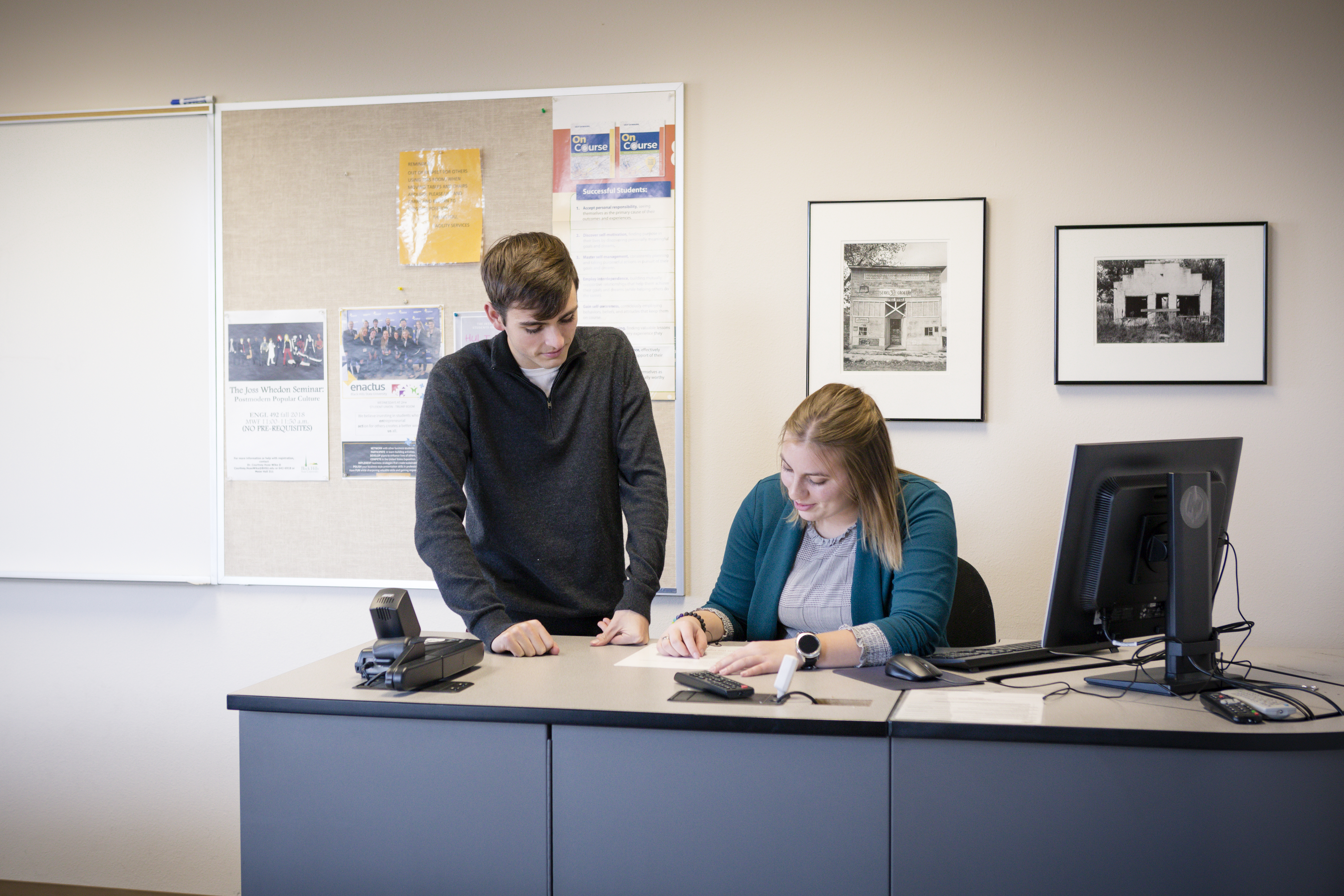 Two Leadership students working together on a project, one standing by the desk and one sitting at the desk.