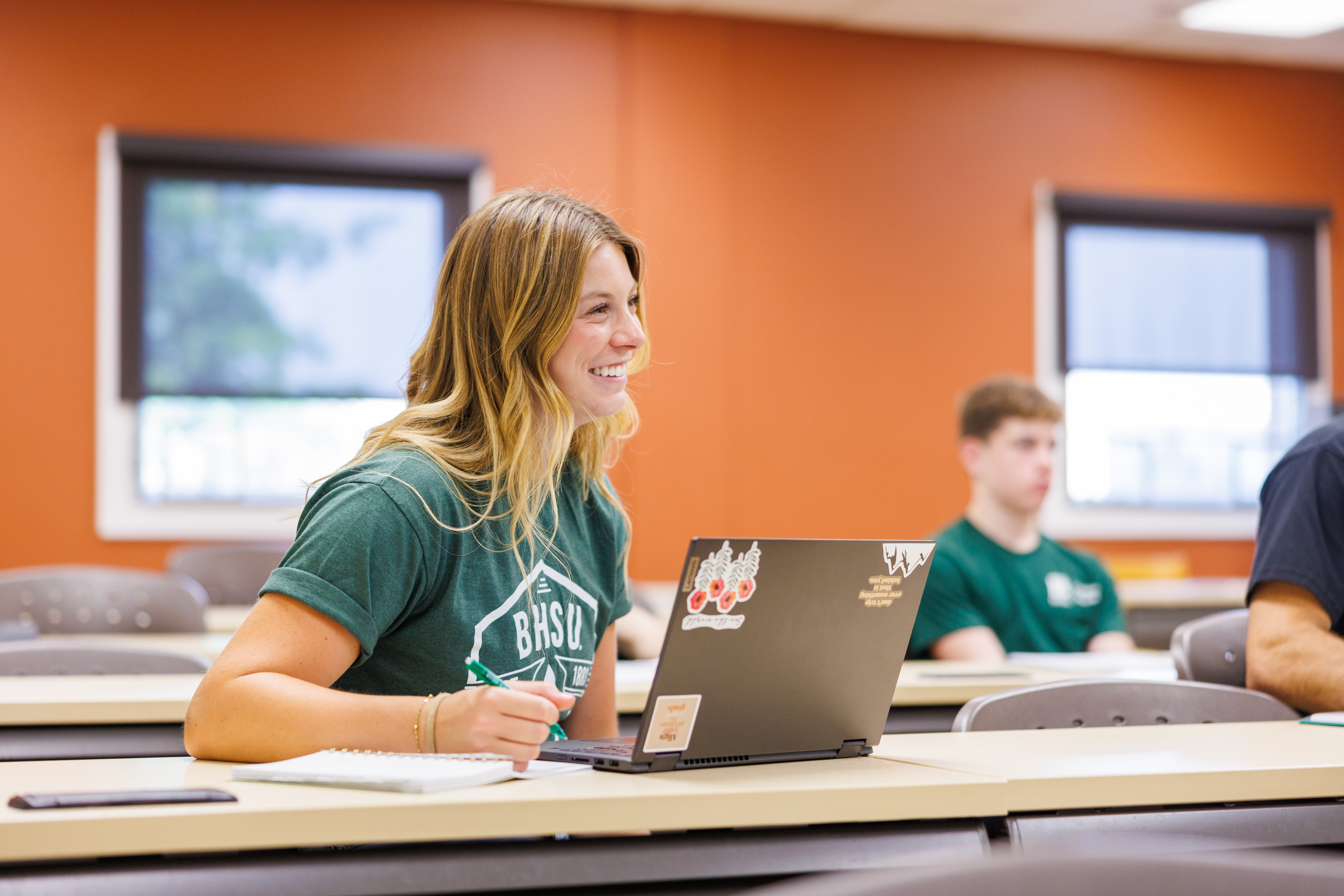 A female BHSU Strategic Leadership student takes notes in a classroom with another student in the background.