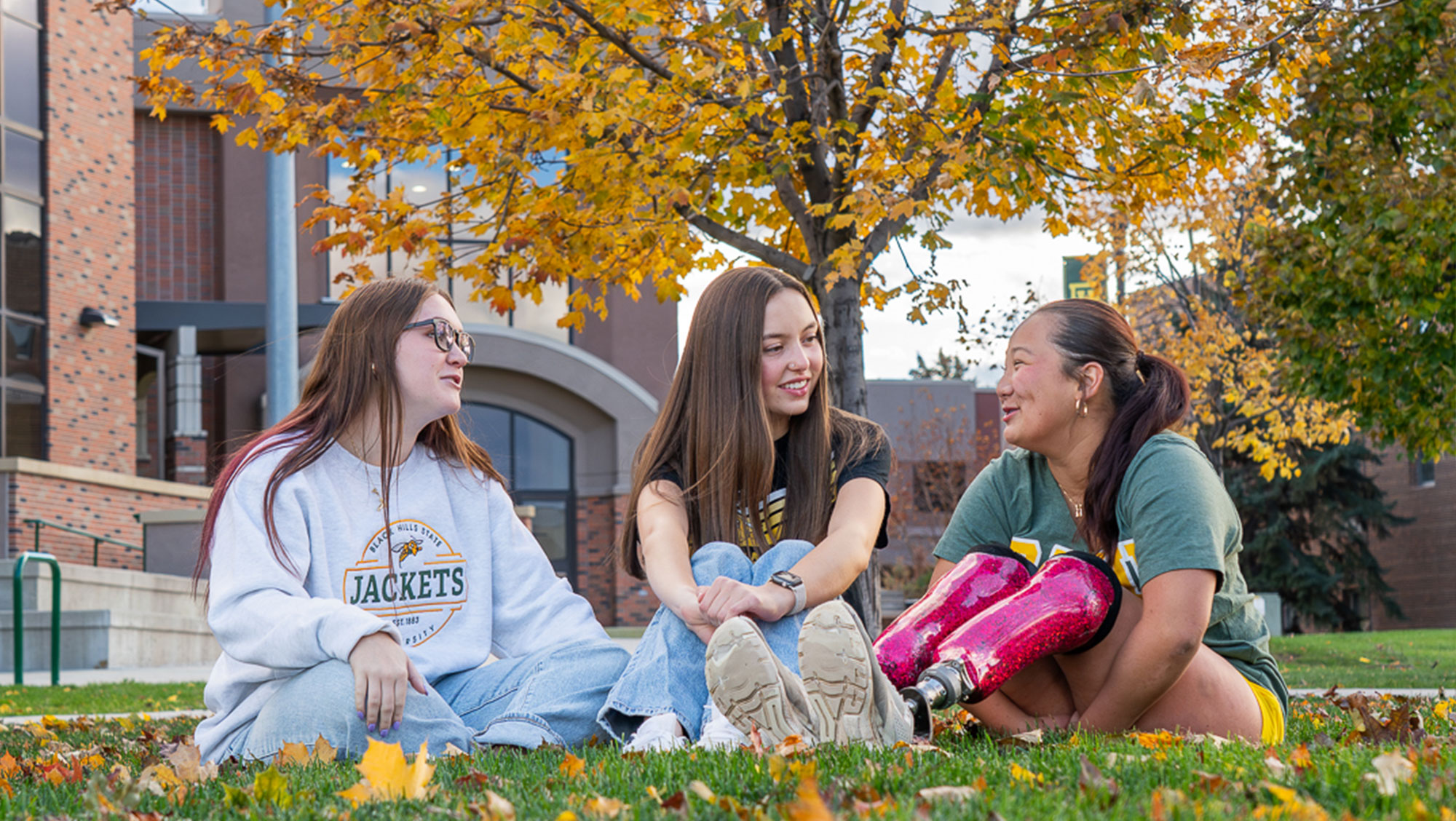 Three BHSU students sitting on a grassy area, chatting and laughing, with autumn trees and a brick building in the background.