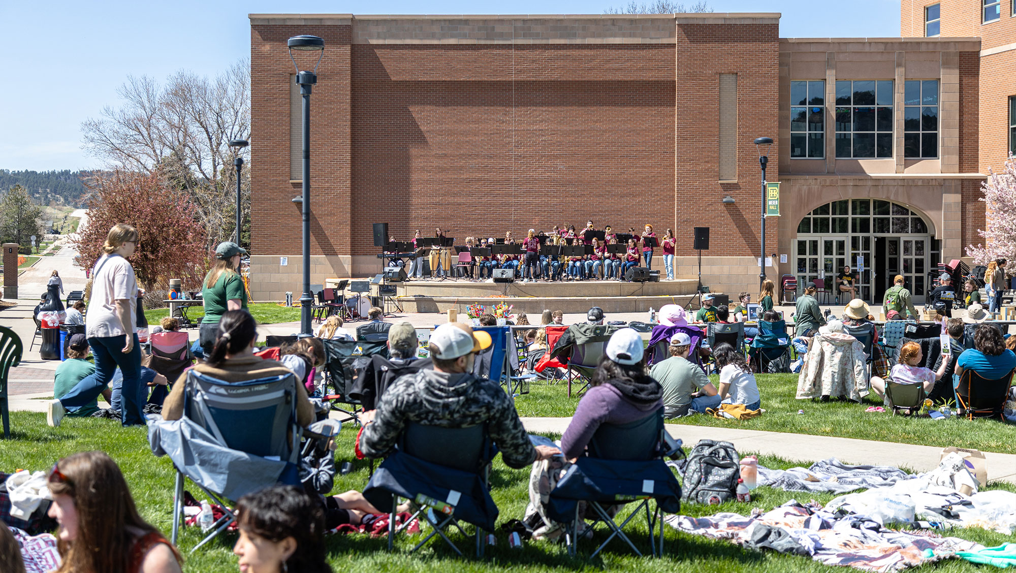 several people sit on blankets and camp chairs on the campus green at BHSU. They are listening to a music group perform on the outdoor stage by Meier Hall on a sunny day.