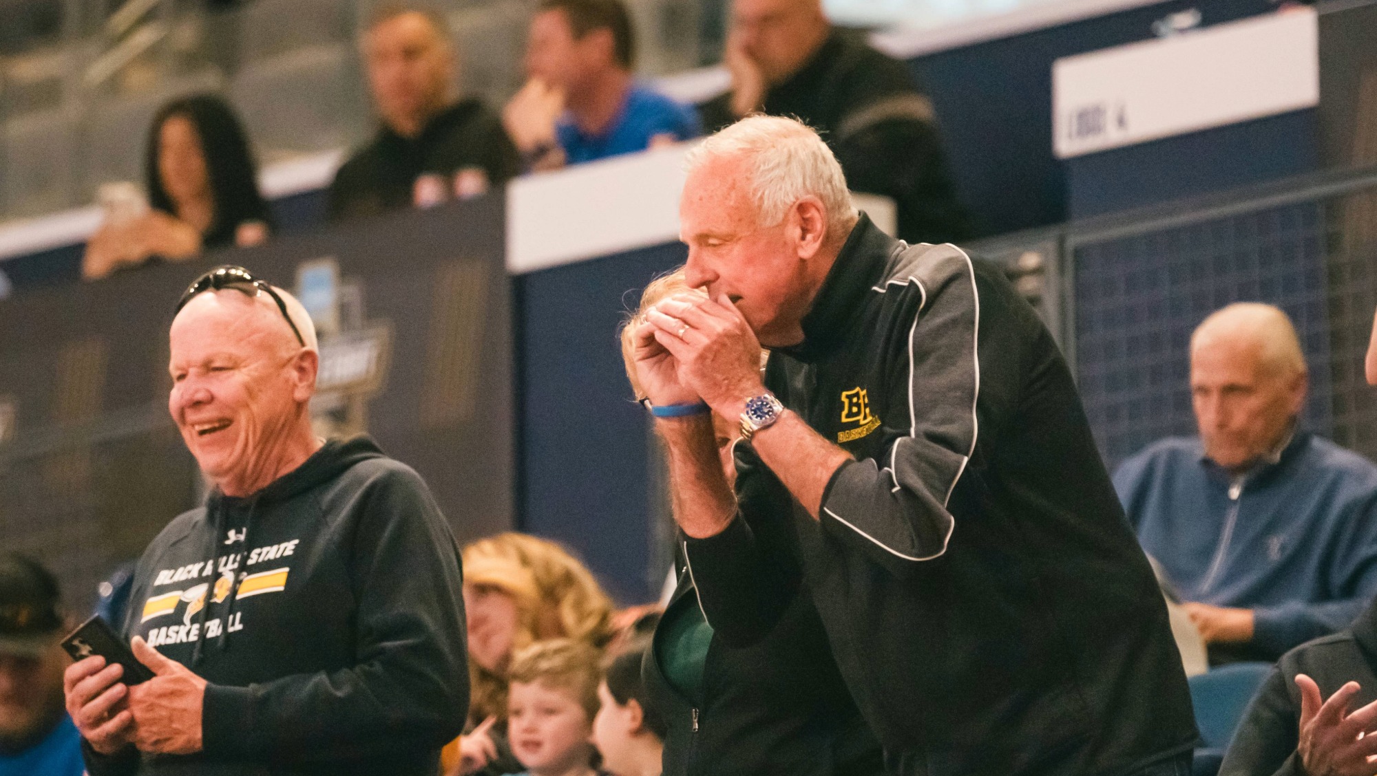 One older man cups his hands around his mouth, raising his voice as he calls out in support of the basketball players on the court. A second man stands in the back watching the game take place. 