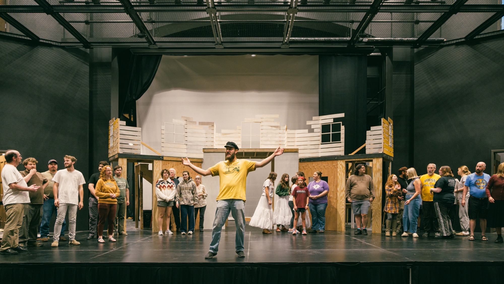 A man stands in the center of the stage arms spread wide while singing with a cast of people surrounding him in the background