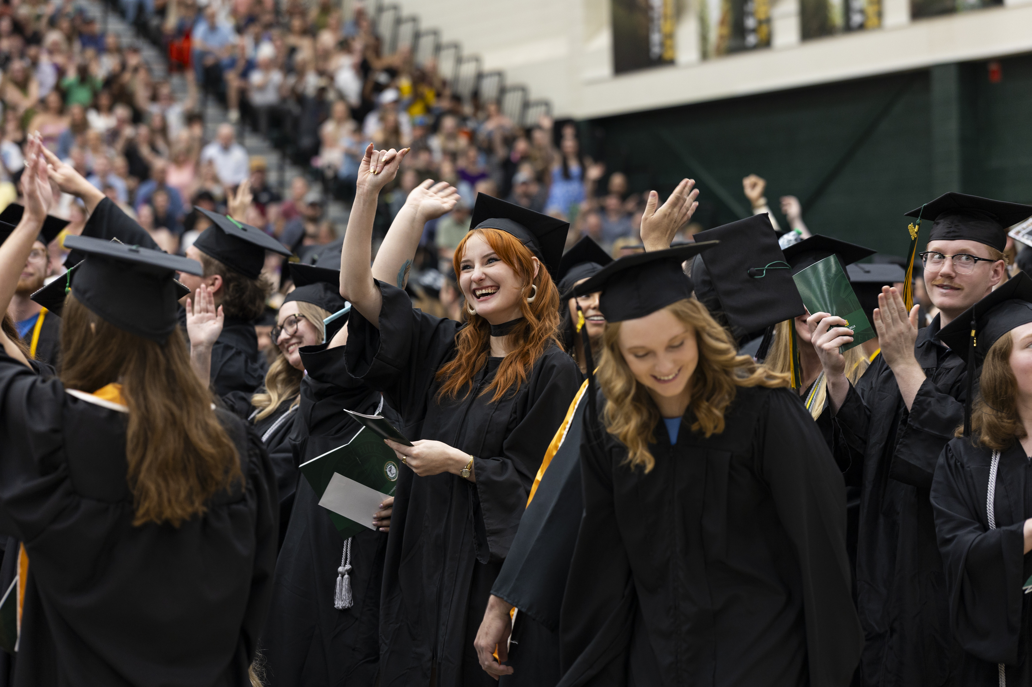 Graduates in caps and gowns cheer and smile during a crowded indoor commencement ceremony.